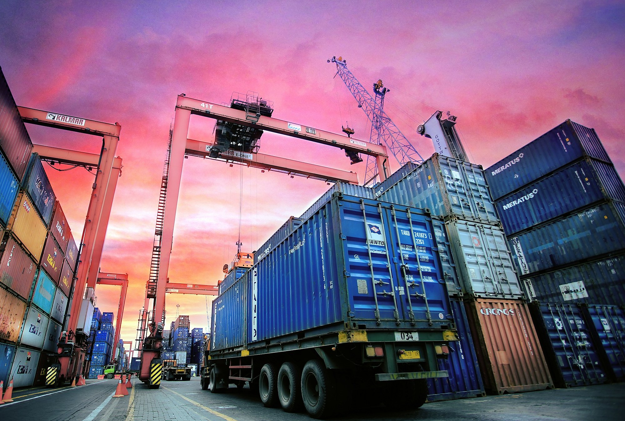 Stacked shipping containers and gantry cranes at a busy port terminal under a vivid sunset sky.