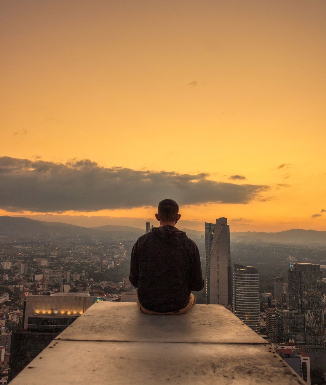 Person seated on a high vantage point looking out over a city skyline at golden hour.