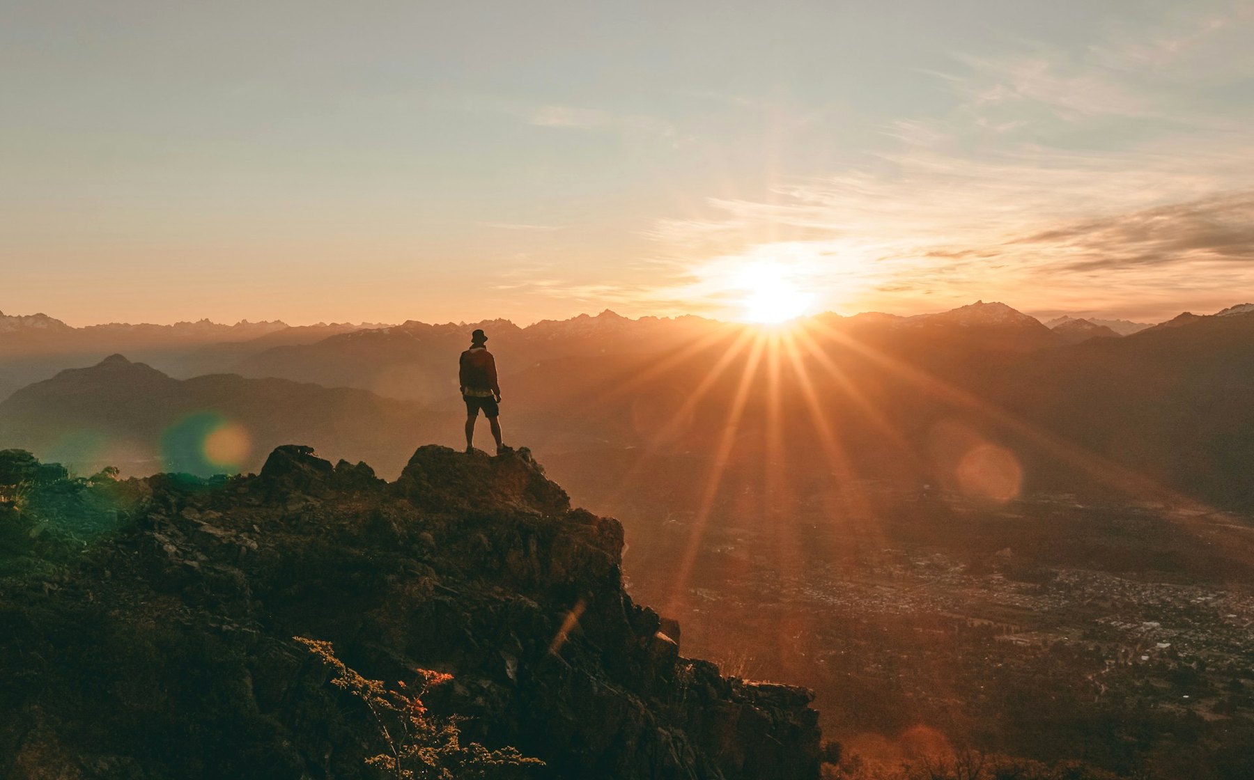 Person standing on a rocky mountain peak at sunset, looking out over a mountain range.